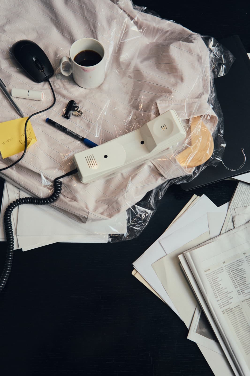 Scrivania in disordine dall'alto: cornetta con filo, tazza di caffè e mouse su una camicia avvolta nella plastica, accanto a fogli sparsi
