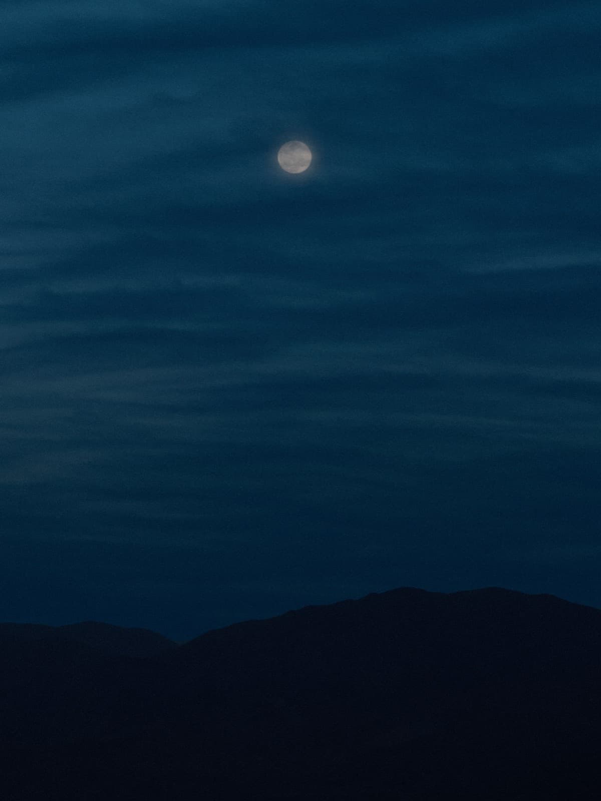 Luna piena che brilla attraverso sottili striature di nuvole, alta sopra la sagoma di una cresta montuosa
