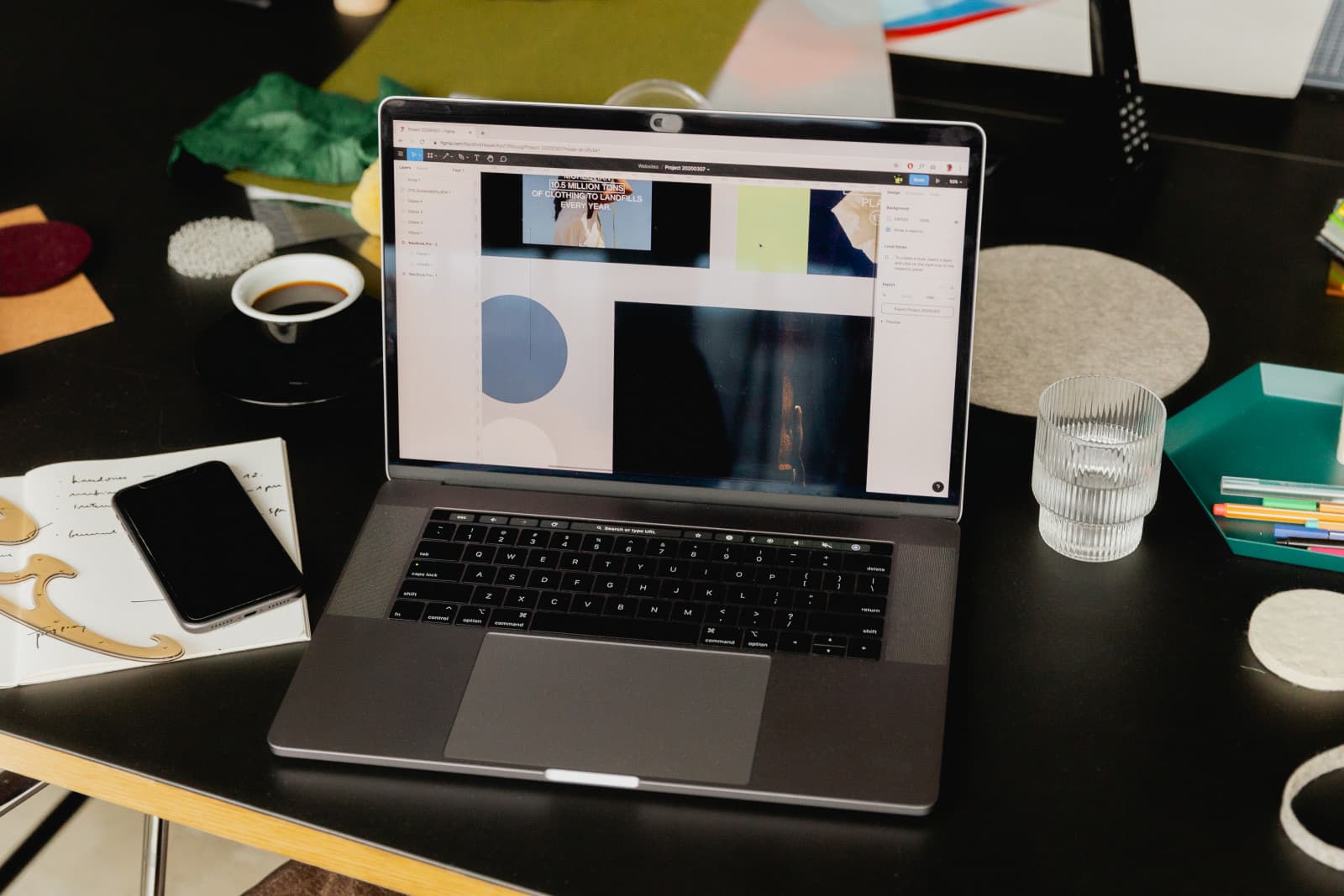 Laptop aperto su un'app di grafica con riquadri di immagini, accanto a un quaderno di appunti a mano, telefono, tazzina e bicchiere d'acqua